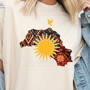 May include: A cream-coloured t-shirt with a colourful map design, a yellow sunburst, and a small bird. The word "Natural" is printed on a white rectangle. The shirt is from the A&Y Apparel collection.