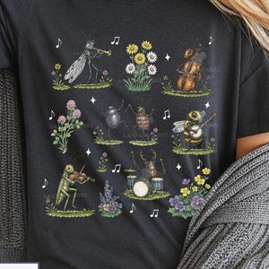 Peut inclure: Un t-shirt gris foncé orné d'une impression graphique représentant divers insectes jouant des instruments de musique, dont une sauterelle au violon, un scarabée à la batterie et une abeille au banjo, entourés de petites illustrations florales.