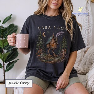 May include: A dark grey t-shirt featuring a graphic print of a house on chicken legs in a forest setting with the text BABA YAGA above the house. The design includes a crescent moon, stars, and smoke rising from the chimney of the house.