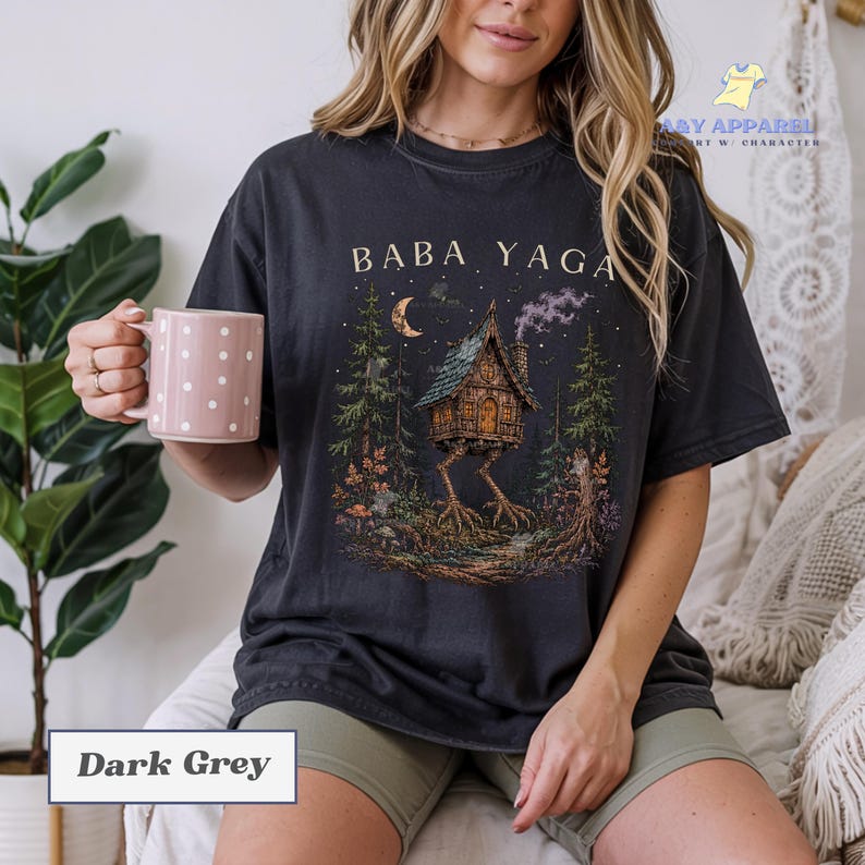 May include: A dark grey t-shirt featuring a graphic print of a house on chicken legs in a forest setting with the text BABA YAGA above the house. The design includes a crescent moon, stars, and smoke rising from the chimney of the house.