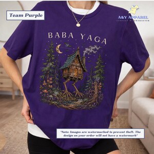 May include: A purple short-sleeve t-shirt featuring a graphic print of a house on chicken legs in a forest setting. The text BABA YAGA is printed above the house. The design includes a crescent moon, trees, and smoke rising from the chimney.