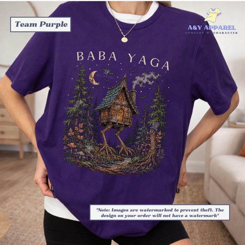 May include: A purple short-sleeve t-shirt featuring a graphic print of a house on chicken legs in a forest setting. The text BABA YAGA is printed above the house. The design includes a crescent moon, trees, and smoke rising from the chimney.