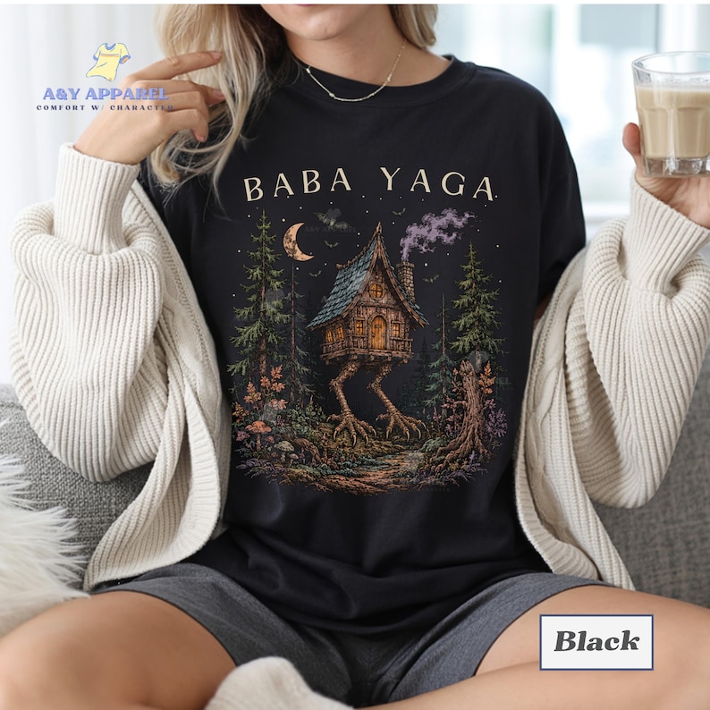 May include: A black t-shirt featuring a graphic print of a house on chicken legs in a forest setting with the text BABA YAGA above it. The illustration includes a crescent moon, pine trees, and smoke rising from the chimney of the wooden house.