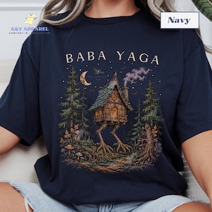 May include: A navy blue t-shirt featuring a graphic print of a wooden hut on chicken legs in a forest setting. The text BABA YAGA is printed above the hut, which is surrounded by pine trees, a crescent moon, and small bats under a starry sky.