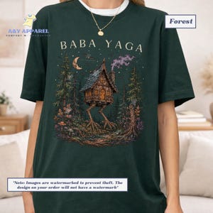 May include: A dark forest green t-shirt featuring a graphic print of a wooden hut on chicken legs in a forest setting. The text BABA YAGA is printed above the hut, which is surrounded by trees, a crescent moon, and smoke in the night sky.