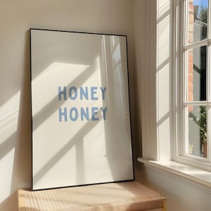 Puede incluir: Un cuadro enmarcado con las palabras "HONEY HONEY" en azul claro sobre un fondo crema. El marco negro está colocado sobre un banco tejido cerca de una ventana, con sombras de luz solar.