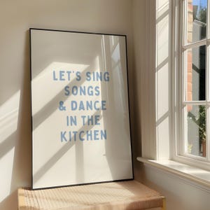 May include: A framed art print with the text "LET'S SING SONGS & DANCE IN THE KITCHEN" in light blue lettering. The print is displayed against a neutral background, with sunlight streaming in from a nearby window.