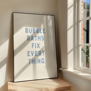 Könnte beinhalten: Ein gerahmter Druck mit den Worten "BUBBLE BATHS FIX EVERYTHING" in hellblauen Buchstaben auf cremefarbenem Hintergrund. Der schwarze Rahmen steht auf einer gewebten Bank in der Nähe eines sonnigen Fensters.