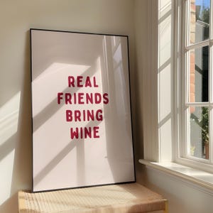 以下が含まれることがあります： 白い背景に、バーガンディ色の文字で「REAL FRIENDS BRING WINE」と書かれたフレーム入りのアートプリント。 黒いフレームは、窓の近くの織り込まれたベンチの上に置かれ、日光が影を作り出しています。