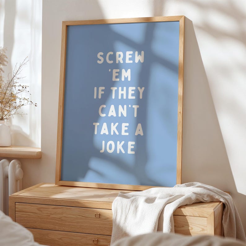 May include: A framed poster with the text "SCREW 'EM IF THEY CAN'T TAKE A JOKE" in white letters on a light blue background. The poster is in a light wood frame and rests on a wooden chest of drawers.