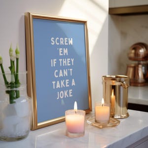 May include: A framed blue print with the text "Screw 'em if they can't take a joke" is displayed on a white surface. Two lit candles and a gold-coloured bucket are next to the frame. A glass vase with white flowers is on the left.