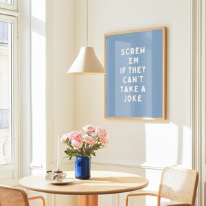 May include: A framed art print with a light blue background and the text "SCREW 'EM IF THEY CAN'T TAKE A JOKE" in white. The print is in a light wood frame. A round wooden table with a blue vase of pink flowers and a teacup is in front of the print.