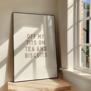 Könnte beinhalten: Ein gerahmter Druck mit dem Text "OFF MY TITS ON TEA AND BISCUITS" in einer neutralen Schriftart. Der Druck wird auf einer gewebten Oberfläche in der Nähe eines Fensters mit Schatten durch Sonnenlicht angezeigt.