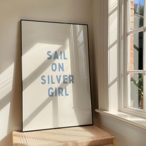 Puede incluir: Una impresión artística enmarcada con las palabras "SAIL ON SILVER GIRL" en letras azul claro sobre un fondo blanco. El marco negro se encuentra sobre un banco tejido cerca de una ventana soleada.