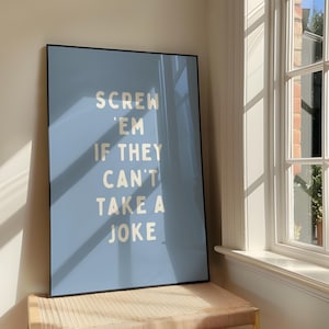 May include: A framed poster with a pale blue background and the text "SCREW 'EM IF THEY CAN'T TAKE A JOKE" in a bold, white font. The poster is leaning against a wall near a window, with sunlight casting shadows.