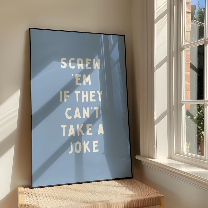 May include: A framed poster with a pale blue background and the text "SCREW 'EM IF THEY CAN'T TAKE A JOKE" in a bold, white font. The poster is leaning against a wall near a window, with sunlight casting shadows.