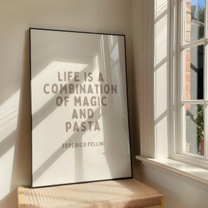 Op de afbeelding: Een ingelijst print met een witte achtergrond en een zwarte lijst. De print bevat een citaat in bruine tekst dat luidt: "Het leven is een combinatie van magie en pasta." Het citaat wordt toegeschreven aan Federico Fellini.