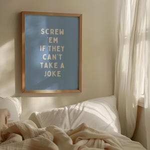 May include: A framed art print with the text "SCREW 'EM IF THEY CAN'T TAKE A JOKE" in a light colour on a blue background. The frame is a light wood colour. The print is hanging above a bed with white pillows and a cream-coloured blanket.