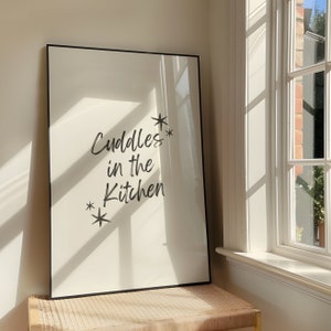 Puede incluir: Una impresión enmarcada en negro con el texto "Cuddles in the Kitchen" en letras blancas con dos pequeñas estrellas a cada lado del texto.