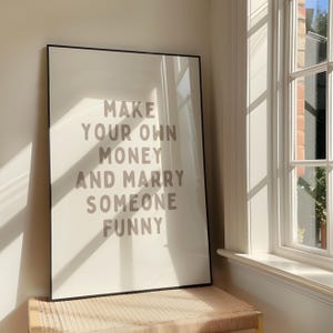 Könnte beinhalten: Ein gerahmter Kunstdruck mit dem Text "MAKE YOUR OWN MONEY AND MARRY SOMEONE FUNNY" in taupefarbenen Buchstaben auf cremefarbenem Hintergrund. Der schwarze Rahmen ruht auf einer gewebten Bank in der Nähe eines Fensters.