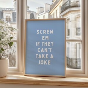 May include: A framed art print with a light blue background and the text "SCREW 'EM IF THEY CAN'T TAKE A JOKE" in white. The frame is made of light-coloured wood. The print is displayed on a windowsill.