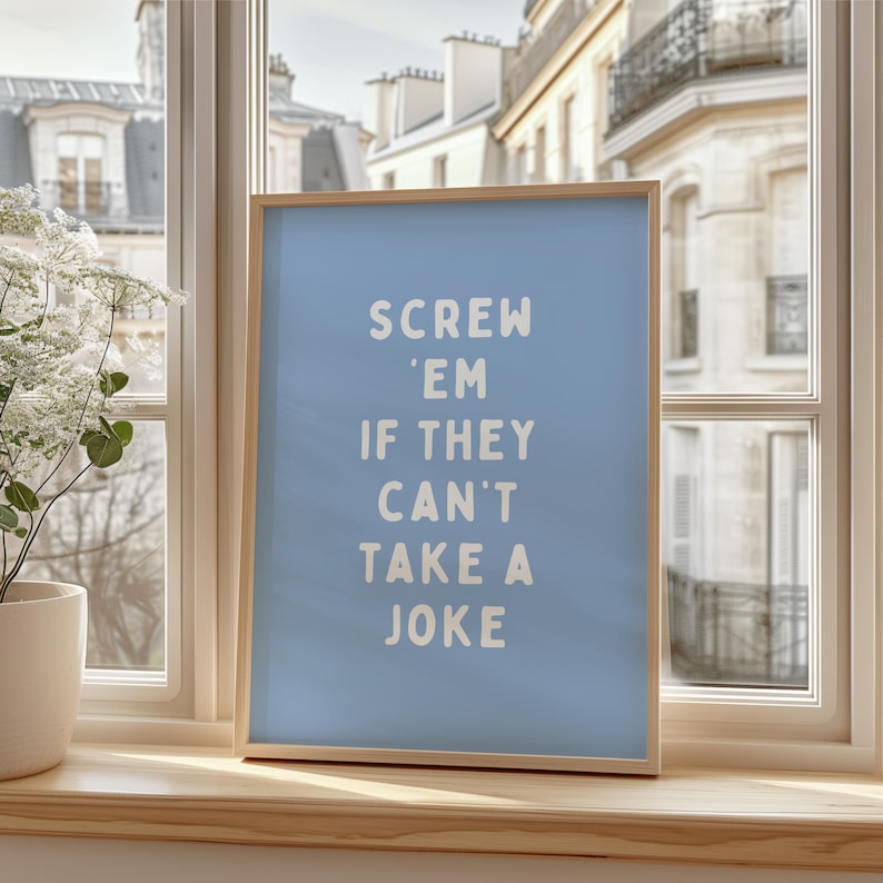 May include: A framed art print with a light blue background and the text "SCREW 'EM IF THEY CAN'T TAKE A JOKE" in white. The frame is made of light-coloured wood. The print is displayed on a windowsill.