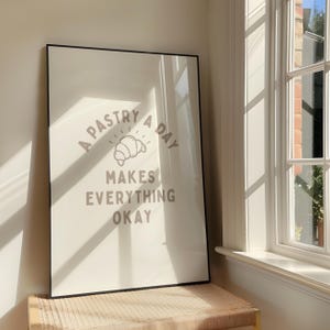 Może przedstawiać: Oprawiony wydruk z napisem "A pastry a day makes everything okay" w kolorze jasnoszarym. Wydruk przedstawia ilustrację rogalika. Rama jest czarna, a wydruk opiera się o ścianę w pobliżu okna.