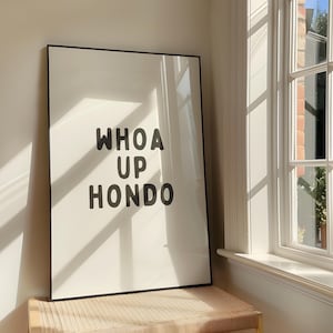 May include: A framed art print featuring the text "WHOA UP HONDO" in black, bold letters on a white background. The print is displayed indoors, near a window. The frame is black.