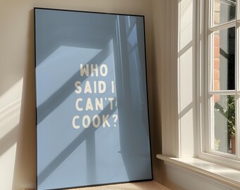 Puede incluir: Un póster enmarcado con un fondo azul claro y el texto "WHO SAID I CAN'T COOK?" en una fuente en negrita, blanquecina. El póster está apoyado contra una pared cerca de una ventana, con sombras de la luz solar.