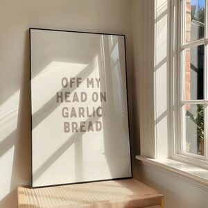 May include: A framed art print with the text "OFF MY HEAD ON GARLIC BREAD" in a light brown font. The print is in a black frame and is leaning against a wall near a window.