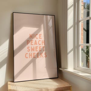 May include: A framed art print with the words "NICE PEACH SWEET CHEEKS" in peach-colored letters on a light pink background. The print is in a black frame and is leaning against a wall near a window.