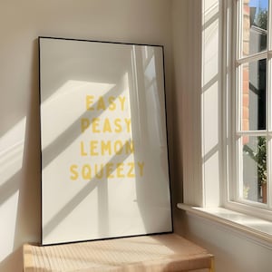 Może przedstawiać: Wydruk w ramie z tekstem "EASY PEASY LEMON SQUEEZY" w żółtych literach na białym tle. Wydruk jest w czarnej ramie i opiera się o ścianę.