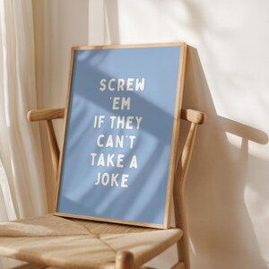 May include: A framed poster with a pale blue background and the text "SCREW 'EM IF THEY CAN'T TAKE A JOKE" in white. The poster is in a light wood frame and is on a wooden chair with a woven seat.