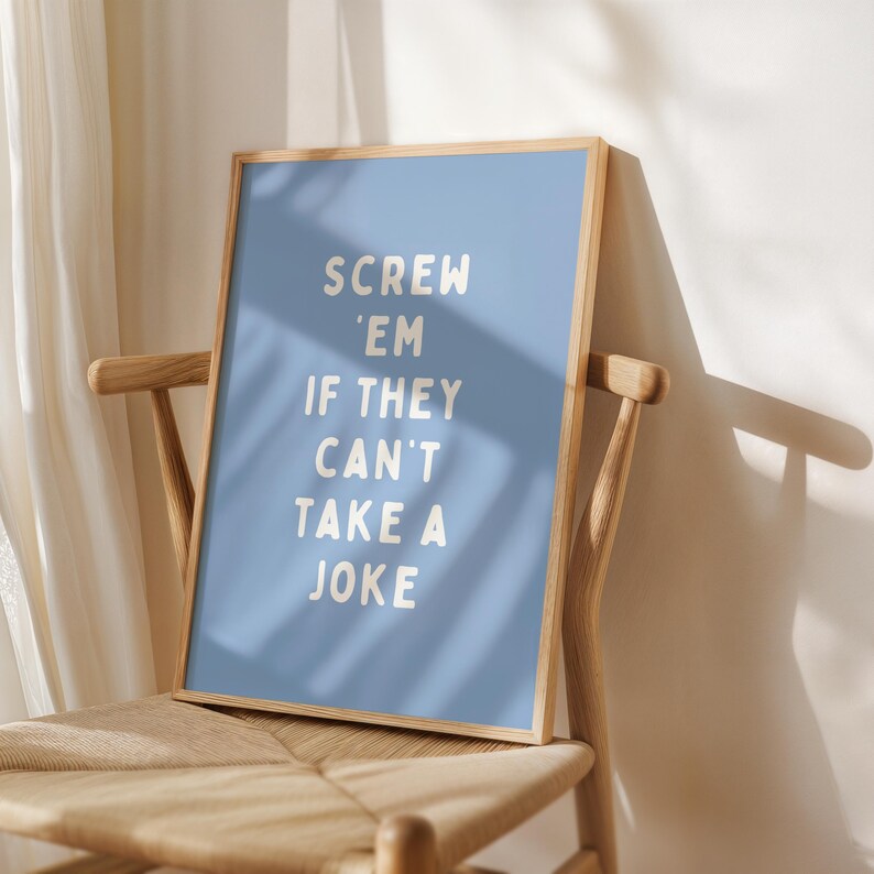 May include: A framed poster with a pale blue background and the text "SCREW 'EM IF THEY CAN'T TAKE A JOKE" in white. The poster is in a light wood frame and is on a wooden chair with a woven seat.
