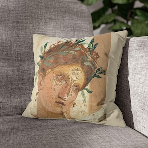 Op de afbeelding: Vierkant sierkussen met een klassiek portret van een persoon met een lauwerkrans. Het kunstwerk heeft een vervaagde, verouderde uitstraling met gedempte tinten beige, bruin en groen. Het kussen ligt op een grijze gestoffeerde bank.
