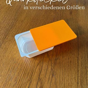 Könnte beinhalten: Ein rechteckiger weißer Kunststoffbehälter mit einem einfarbigen orangefarbenen Deckel. Der Text "Quarkdeckel in verschiedenen Größen" ist oben im Bild zu sehen und weist auf einen Deckel für Milchprodukte in verschiedenen Größen hin.