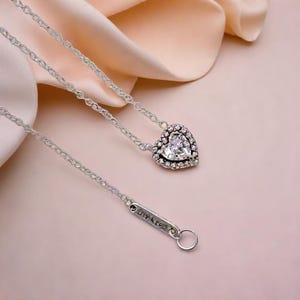 May include: A silver necklace featuring a heart-shaped pendant. The pendant is adorned with clear crystals surrounding a central stone. The delicate chain leads to a clasp with engraved text.