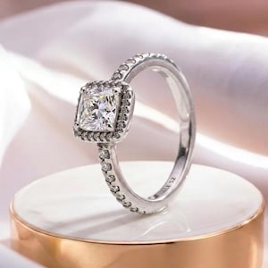 May include: A silver-toned ring featuring a central cushion-cut clear stone surrounded by a halo of smaller clear stones. The band is also set with small clear stones along its sides. The interior of the band is engraved with the text ALE 52.