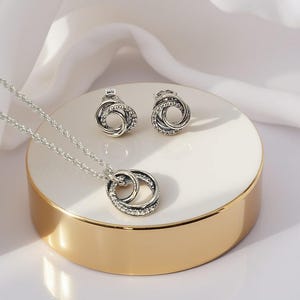 May include: A silver jewellery set comprising a necklace and stud earrings. The necklace features a pendant with two interlocking circles, and the earrings have a similar design. Both are adorned with small, clear crystals. The set is presented on a white and gold platform.