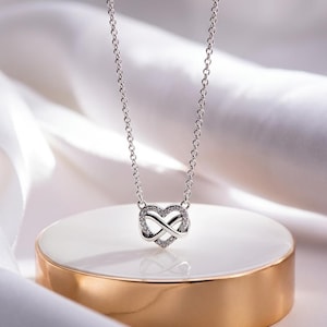 May include: A silver-coloured necklace featuring a pendant that combines a heart shape with an infinity symbol. The pendant is embellished with small, clear, sparkling stones along the metal frame and hangs from a delicate, thin silver-coloured chain.