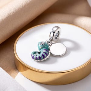Può includere: Un ciondolo in argento a forma di piccolo camaleonte arrotolato con dettagli in smalto color ottanio e viola. Il ciondolo include un disco circolare in argento lucido fissato a una contromaglia, adatto per bracciali o catenine.