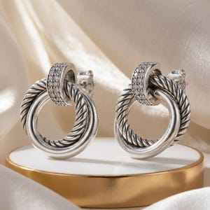 Peut inclure: Une paire de boucles d'oreilles argentées. Chaque boucle d'oreille présente un design à double boucle avec un motif texturé en forme de corde torsadée. La boucle supérieure est ornée de petites pierres claires scintillantes. Les boucles sont présentées sur une surface blanche et dorée.