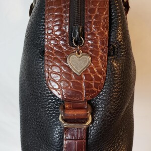 Puede incluir: Bolso de mano de cuero negro con detalles en marr&oacute;n con textura de cocodrilo. Cuenta con una cremallera plateada y un charm en forma de coraz&oacute;n. El bolso tiene asas plateadas y un cierre de hebilla. Un accesorio elegante y funcional.