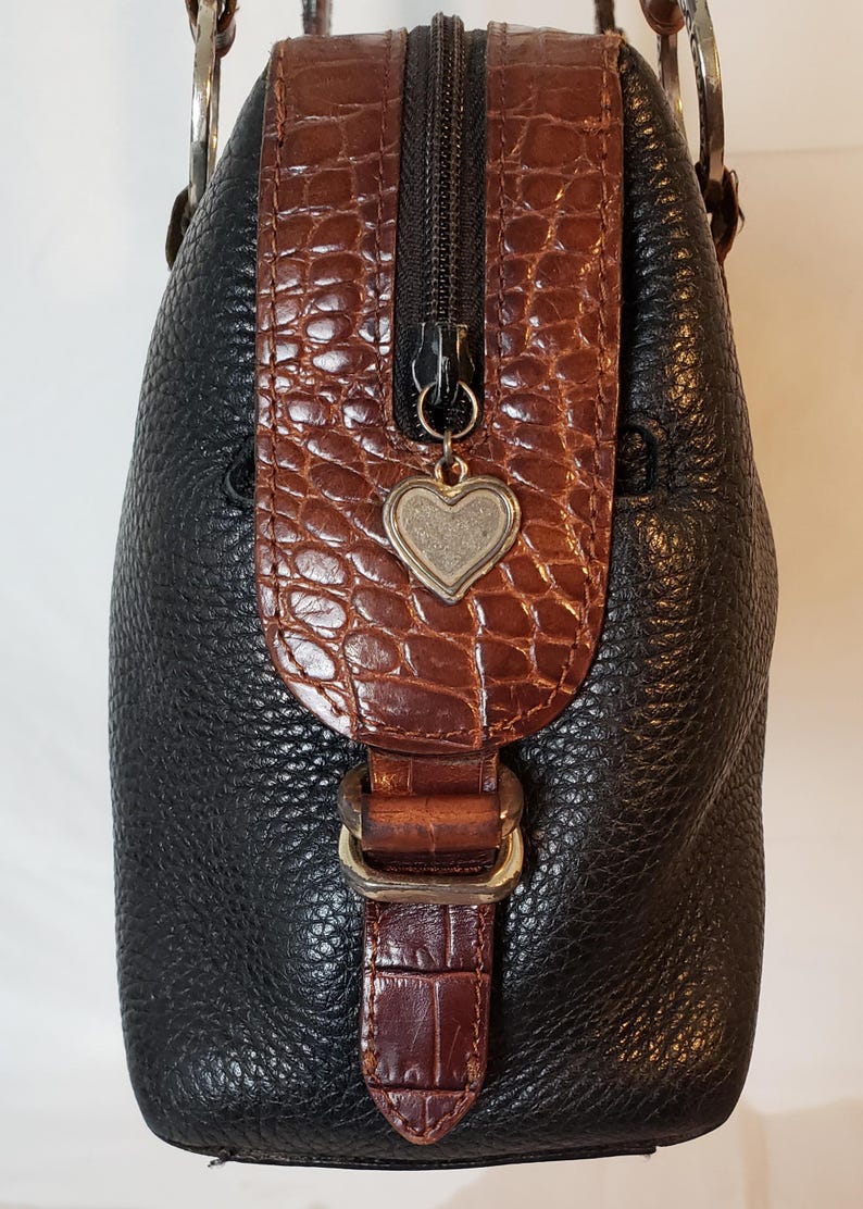Puede incluir: Bolso de mano de cuero negro con detalles en marr&oacute;n con textura de cocodrilo. Cuenta con una cremallera plateada y un charm en forma de coraz&oacute;n. El bolso tiene asas plateadas y un cierre de hebilla. Un accesorio elegante y funcional.
