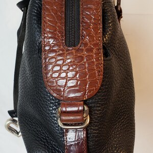 Puede incluir: Bolso de mano de cuero negro con detalles marrones con textura de cocodrilo. El bolso tiene una cremallera superior, un cierre de hebilla y herrajes plateados. El dise&ntilde;o combina cuero liso y texturizado.