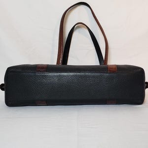 Puede incluir: Bolso tote de cuero negro con detalles y asas marrones. El bolso presenta un exterior texturizado y una forma rectangular. Las asas son marrones y el bolso tiene detalles marrones en los laterales. Dise&ntilde;ado para transportar art&iacute;culos.