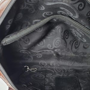 Puede incluir: Bolso de mano de cuero marr&oacute;n y negro con herrajes plateados. El interior presenta un forro de tela negra, un bolsillo con cremallera y un llavero. El bolso tiene cierre de cremallera superior y dos asas.