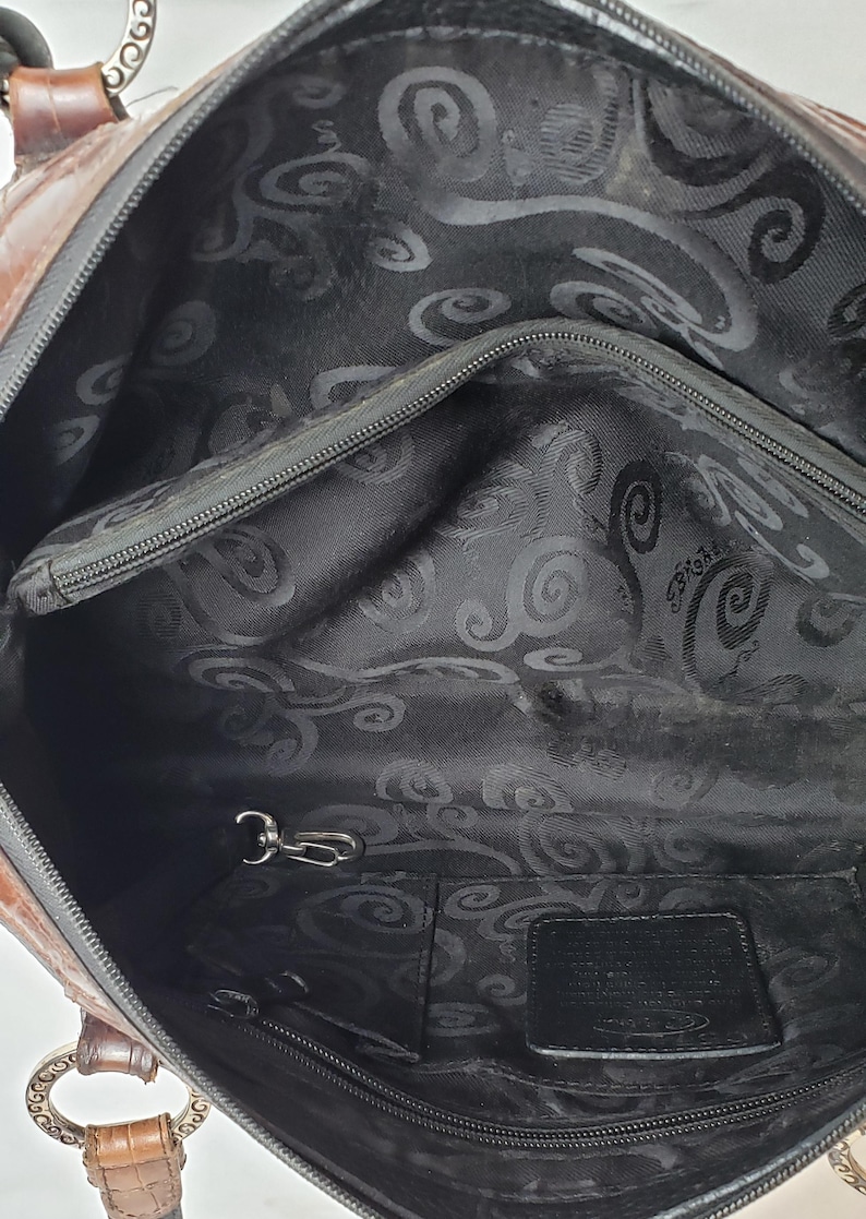 Puede incluir: Bolso de mano de cuero marr&oacute;n y negro con herrajes plateados. El interior presenta un forro de tela negra, un bolsillo con cremallera y un llavero. El bolso tiene cierre de cremallera superior y dos asas.