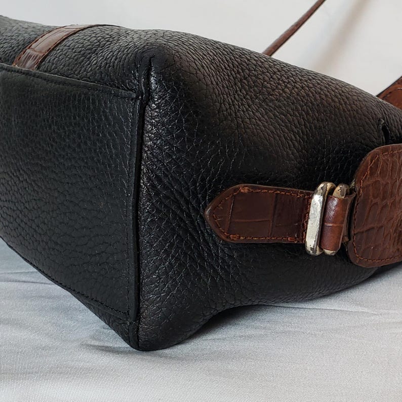 Puede incluir: Bolso de mano de cuero negro con detalles de cuero marr&oacute;n con relieve de cocodrilo. El bolso tiene forma rectangular con una superficie texturizada y una hebilla plateada. Con correa para el hombro.