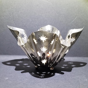 May include: A silver-colored, star-shaped candle holder with a ruffled edge. The candle holder has star cutouts that cast star-shaped shadows. The candle holder sits on three small, gold-colored feet.
